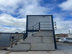 Escalier en blocs béton gris avec rampe métallique noire menant à une structure modulaire blanche, dans un environnement industriel extérieur.